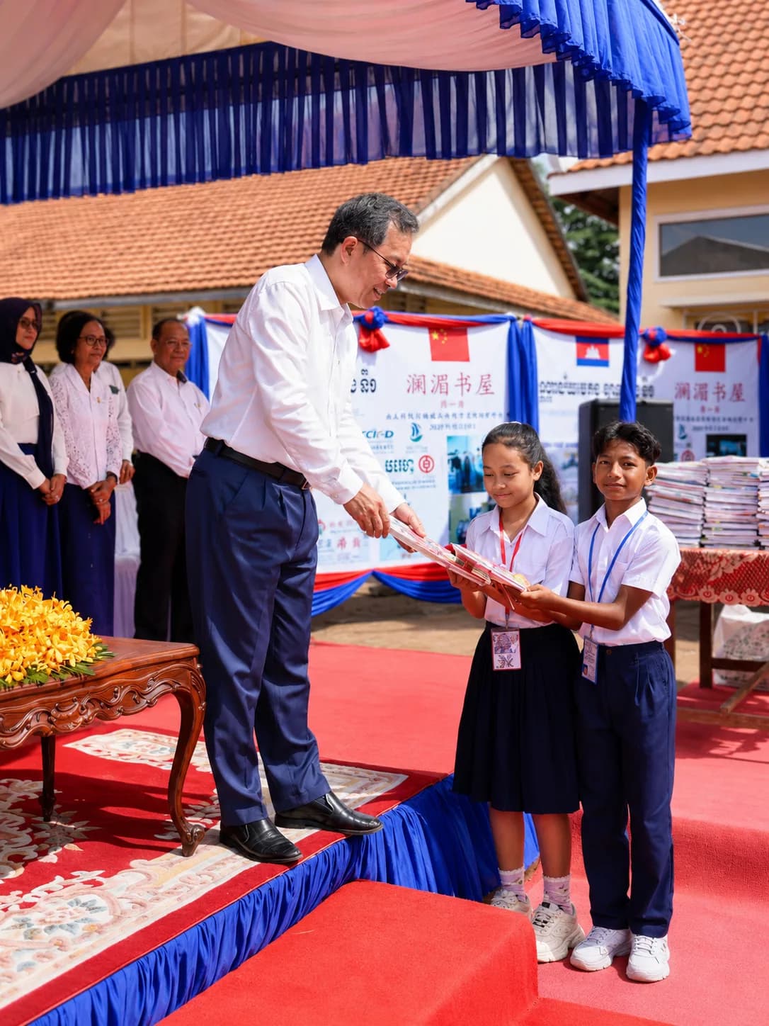 A man hands books to two students during a school ceremony, with officials and stacked books in the background.