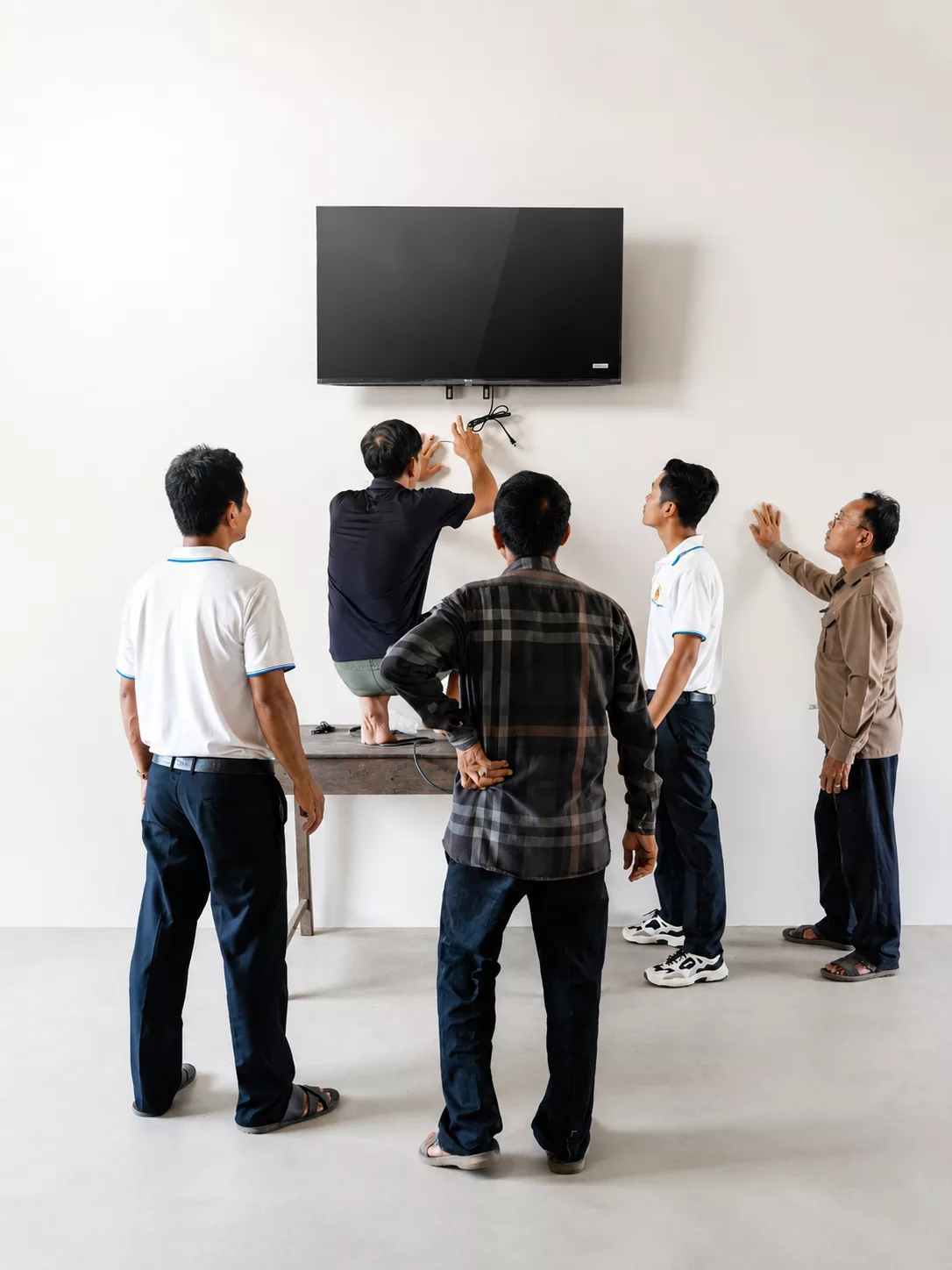 Five men collaboratively installing a wall-mounted TV in a clean minimalist room, with one person adjusting the cables while the others observe and assist.