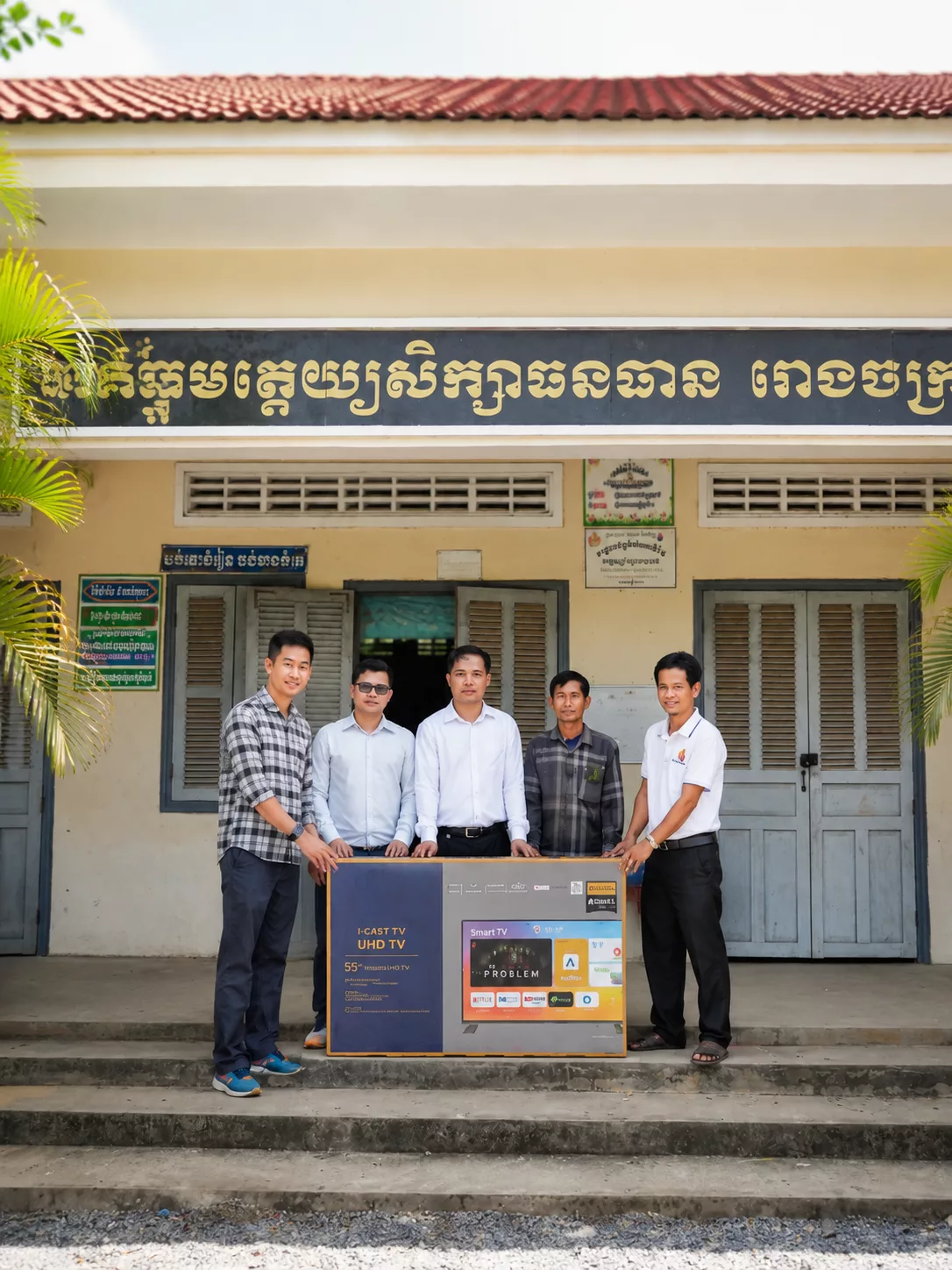A group of five men pose outside a school building while presenting a boxed smart TV donation, with Khmer signage above the entrance.